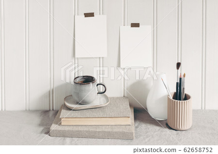Blank paper cards taped on white wooden wall. Stationery mockup scene with paint brushes, pencils in ceramic holder, cup of coffee and books on linen tablecloth. Artistic scene. Creative background Blank paper cards taped on white wooden wall. Stationery mockup scene with paint brushes, pencils in ceramic holder, cup of coffee and books on linen tablecloth. Artistic scene. Creative background 62658752