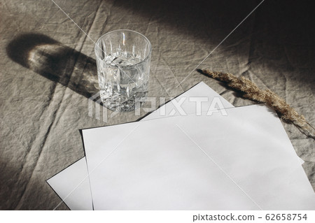Stationery still life scene. Glass of water, dry grass on beige linen table cloth background in sunlight. Blank paper sheets, cards mockup scene with long harsh shadows. High angle view. Stationery still life scene. Glass of water, dry grass on beige linen table cloth background in sunlight. Blank paper sheets, cards mockup scene with long harsh shadows. High angle view. 62658754