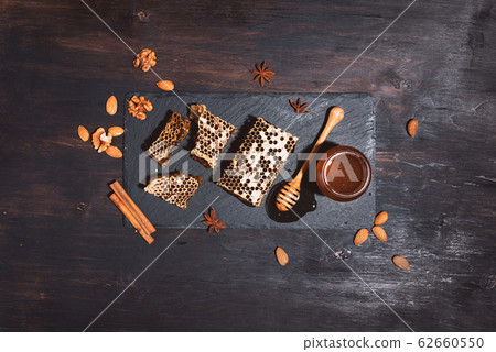 Honeycomb, bee pollen granules, honey jar with wooden dipper, almond nuts on black slate tray over dark background. Copy space. Top view. Autumn harvest concept Honeycomb, bee pollen granules, honey jar with wooden dipper, almond nuts on black slate tray over dark background. Copy space. Top view. Autumn harvest concept 62660550