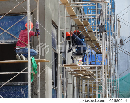 Brick wall plastered by construction workers using the cement plaster. Scaffolding used as temporary staging to work at height. Wearing appropriate safety gear to prevent bad happen. Brick wall plastered by construction workers using the cement plaster. Scaffolding used as temporary staging to work at height. Wearing appropriate safety gear to prevent bad happen. 62680944