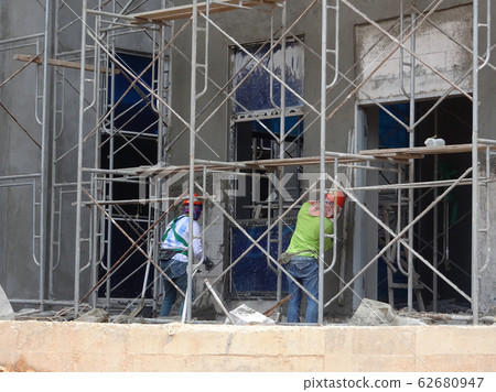Brick wall plastered by construction workers using the cement plaster. Scaffolding used as temporary staging to work at height. Wearing appropriate safety gear to prevent bad happen.   62680947