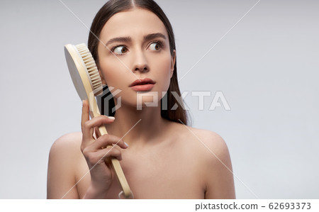 Stunned young woman with massage brush standing against light blue background Stunned young woman with massage brush standing against light blue background 62693373