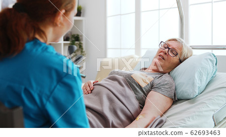 In nursing home old sick lady lying in bed talks with a female nurse 62693463