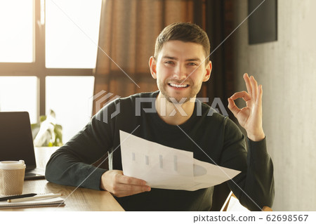 Guy Gesturing Okay Holding Papers With Charts Sitting At Workplace Guy Gesturing Okay Holding Papers With Charts Sitting At Workplace 62698567