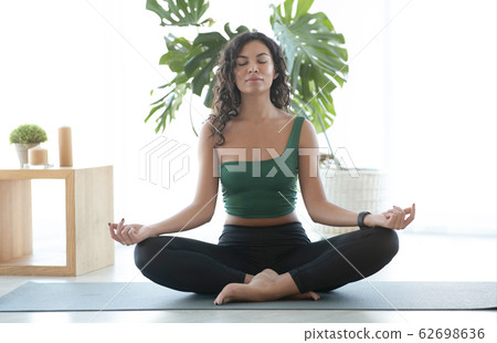 Young Hispanic woman doing meditation in lotus yoga pose at home Young Hispanic woman doing meditation in lotus yoga pose at home 62698636