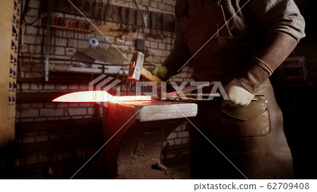 A man blacksmith forging a hot knife blade using a hammer 62709408