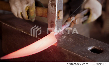 A man blacksmith hitting a hot knife blade with a hammer 62709416