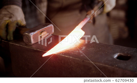 A man blacksmith forging a hot knife blade with a hammer 62709430