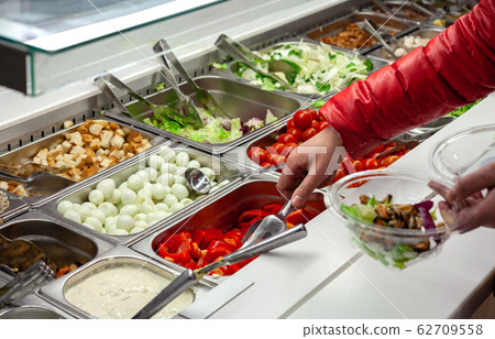 Fresh Salad bar counter with person's hands lifting vegetable into a salad bowl. Fresh Salad bar counter with person's hands lifting vegetable into a salad bowl. 62709558