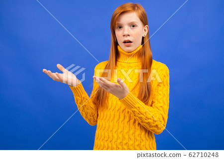 charming european red-haired girl in a yellow sweater waving her arms and indignant on a blue studio charming european red-haired girl in a yellow sweater waving her arms and indignant on a blue studio 62710248