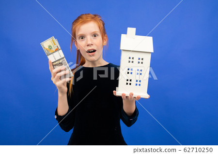 surprised red-haired girl with money and a mock up of a house on a blue background. buying a surprised red-haired girl with money and a mock up of a house on a blue background. buying a 62710250