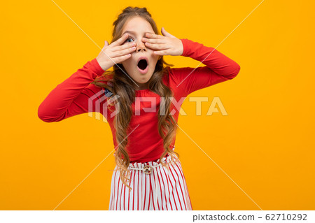 surprised teenager girl with curly hair in a red blouse and striped trousers covers her hands with 62710292