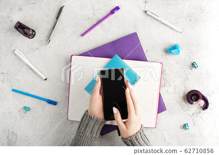 Female hands holding smartphone at workplace with notepads, pens and other office supplies. Women's business concept. Flat lay. 62710856