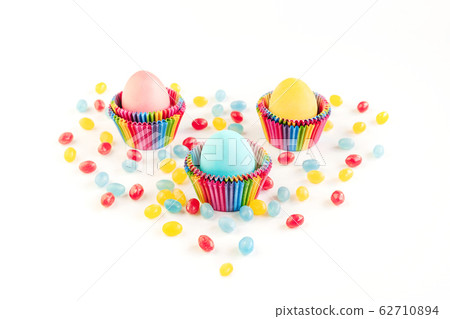 Bright creative Easter decoration of colorful eggs in raibow colored paper forms for cupcakes on white background decorated with jelly candies. Place for text. 62710894