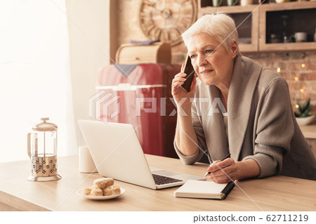 Senior Businesswoman Using Laptop, Talking On Cellphone And Taking Notes 62711219