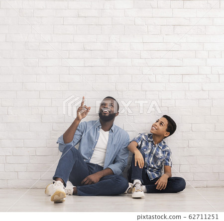 Afro Son And Dad Sitting Near Brick Wall And Pointing Upwards Afro Son And Dad Sitting Near Brick Wall And Pointing Upwards 62711251
