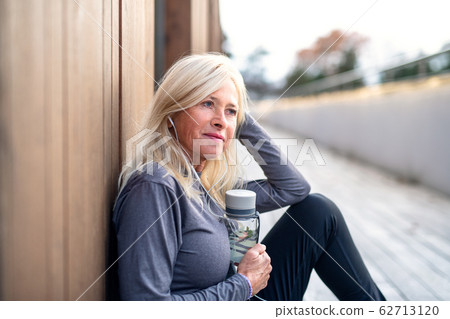 A senior woman with earphones sitting outdoors resting after exercise. A senior woman with earphones sitting outdoors resting after exercise. 62713120