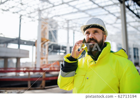 Man engineer standing on construction site, using smartphone. 62713194