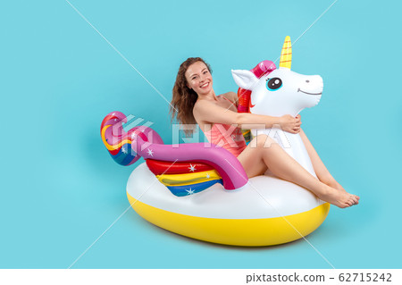 Summer Freestyle. Young woman with freckles isolated on blue sitting on swim ring unicorn shape smiling happy 62715242