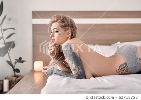 Weekend at Home. Young tattooed woman in lingerie lying on bed at bedroom touching lips playful Weekend at Home. Young tattooed woman in lingerie lying on bed at bedroom touching lips playful 62715258