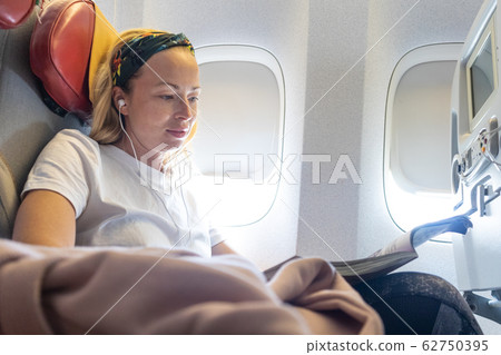 Casual young woman reading magazine and listening to music on airplane. Female traveler reading seated in passanger cabin. 62750395
