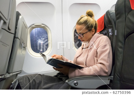 Casual young woman reading magazine and listening to music on airplane. Female traveler reading seated in passanger cabin. 62750396