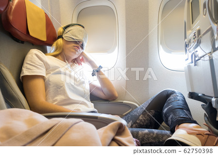 Tired blonde casual caucasian lady napping on uncomfortable seat while traveling by airplane. 62750398
