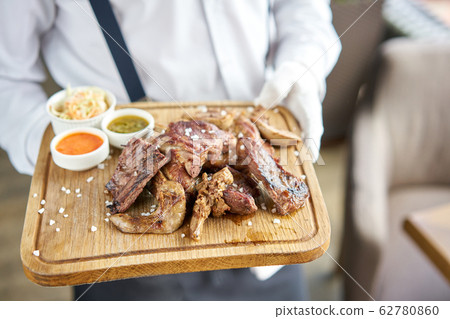 A waiter in a white shirt and apron holds a ready meal in the restaurant. Various grilled meat set with Pickled cabbage and two sauces. Serving on a wooden Board. 62780860