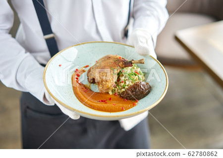 A waiter in a white shirt and apron holds a ready meal in the restaurant.Plate Duck leg su-vide, with bulgur and onion marmalade. 62780862