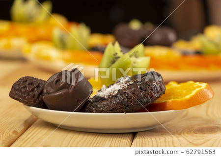 Chocolate candies and cookies sprinkled with coconut flakes lie with chopped fruits on a white saucer on a wooden table. Homemade baking with health benefits. Healthy meal. Delicious vegan snack Chocolate candies and cookies sprinkled with coconut flakes lie with chopped fruits on a white saucer on a wooden table. Homemade baking with health benefits. Healthy meal. Delicious vegan snack 62791563