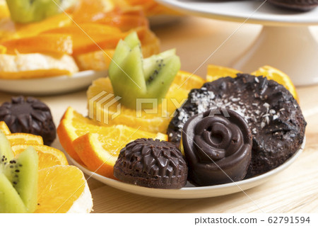 Delicious vegan snack. Chocolate candies and cookies sprinkled with coconut flakes lie with chopped fruits on a white saucer on a wooden table. Homemade baking with health benefits. Healthy meal Delicious vegan snack. Chocolate candies and cookies sprinkled with coconut flakes lie with chopped fruits on a white saucer on a wooden table. Homemade baking with health benefits. Healthy meal 62791594