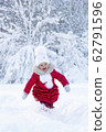 A beautiful little girl in a red coat and white hat with a scarf runs along a snowy path in a winter park. Smile and children's joy on the face. Enjoyment in the winter season. Great holidays. Snow 62791596