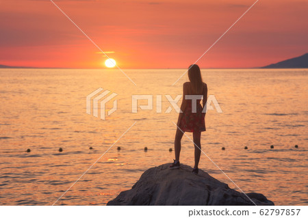 Young woman standing on the rock in the sea against mountains and colorful orange sky at sunset  62797857