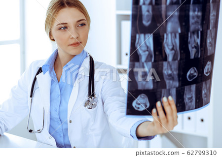 Doctor woman examining x-ray picture near window in hospital. Surgeon or orthopedist at work. Medicine and healthcare concept. Blue colored blouse of a therapist looks good Doctor woman examining x-ray picture near window in hospital. Surgeon or orthopedist at work. Medicine and healthcare concept. Blue colored blouse of a therapist looks good 62799310