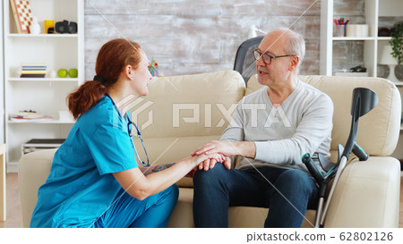 Female nurse talking and taking care of old disabled man sitting on the sofa Female nurse talking and taking care of old disabled man sitting on the sofa 62802126