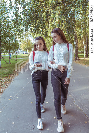 Two girls schoolgirls, walk by hand, returning home after school college. In summer city, background road trees, hand smartphone. Backpacks behind. Casual wear sweater jeans. Social networks online. Two girls schoolgirls, walk by hand, returning home after school college. In summer city, background road trees, hand smartphone. Backpacks behind. Casual wear sweater jeans. Social networks online. 62809682