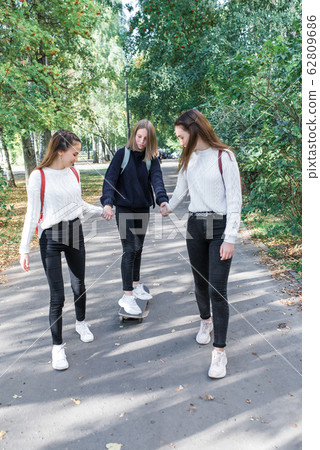 Three teenage girls, in summer in city, ride skateboard, rest break after school weekend. Casual warm clothing sweater jeans. Emotions of joy fun entertainment. Background lawn trees . 62809686