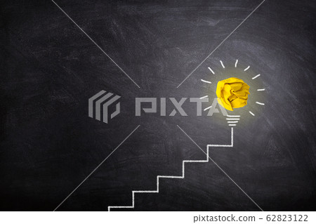 Yellow crumpled paper ball light bulb lighting grow on the top of white chalk ladder handwriting on chalkboard. Yellow crumpled paper ball light bulb lighting grow on the top of white chalk ladder handwriting on chalkboard. 62823122