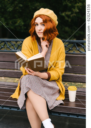 A beautiful girl with a curly hairstyle is sitting on a park bench and reading 62824336