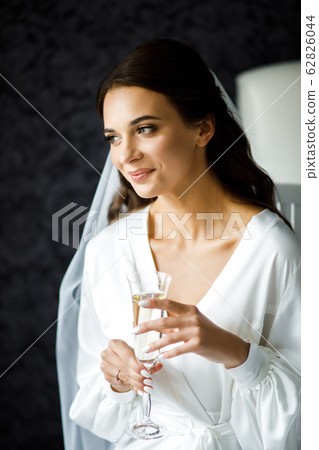 The girl sits on a soft bed, holding a glass with champagne in her hands. Wedding morning of the bride. 62826044