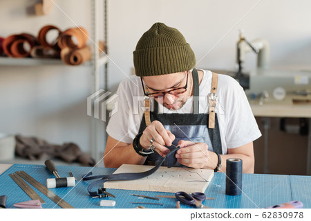 Young creative leatherworker bending over table while sewing new item Young creative leatherworker bending over table while sewing new item 62830978