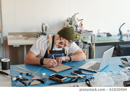 Young leatherworker concentrating upon drawing sketch on page of notepad 62830994