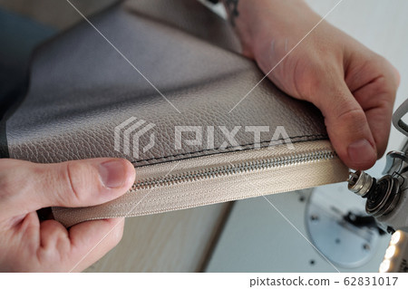 Hands of male leatherworker holding piece of leather with zipper by machine 62831017