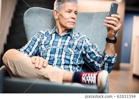 Mature male holding smartphone while resting in armchair 62831394