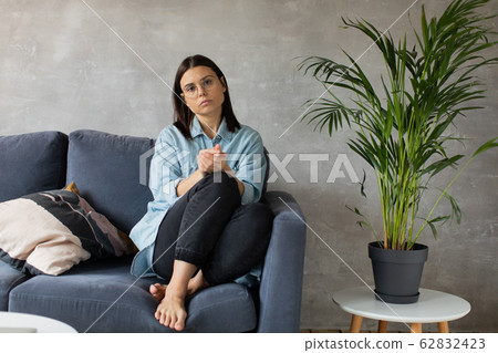 A young Woman is Sitting on the Sofa Looking at the Camera. The Girl Is Worried, Sad. Depression. A young Woman is Sitting on the Sofa Looking at the Camera. The Girl Is Worried, Sad. Depression. 62832423