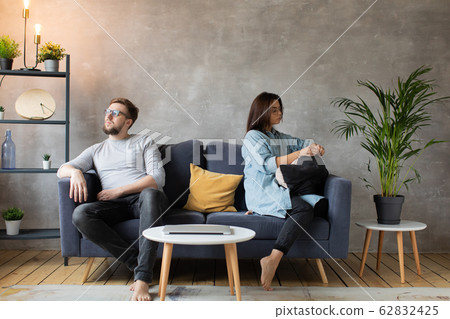 A young Couple are Sitting on the Couch After an Argument at Home. The husband and Wife Sit with their Backs to Each other. A young Couple are Sitting on the Couch After an Argument at Home. The husband and Wife Sit with their Backs to Each other. 62832425
