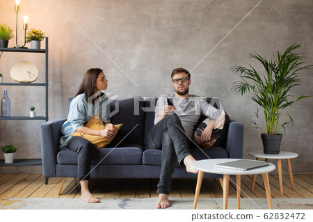 Young Couple Sitting at Home on the Couch. Man Watches TV. Young Woman Calls her Husband, he does not Hear 62832472