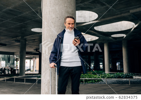 Senior man with rolling suitcase standing outdoors looking at mobile phone Senior man with rolling suitcase standing outdoors looking at mobile phone 62834375