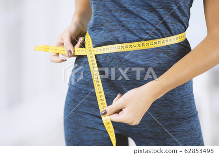 Weight loss concept. Slim woman in sportswear measuring her waist with tape measure indoors, closeup view 62853498