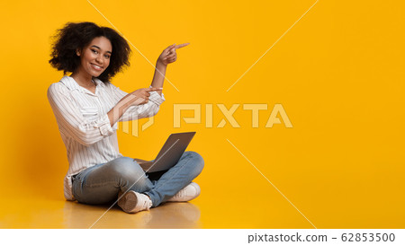 Cheerful Afro Girl Sitting On Floor With Laptop And Pointing Aside Cheerful Afro Girl Sitting On Floor With Laptop And Pointing Aside 62853500